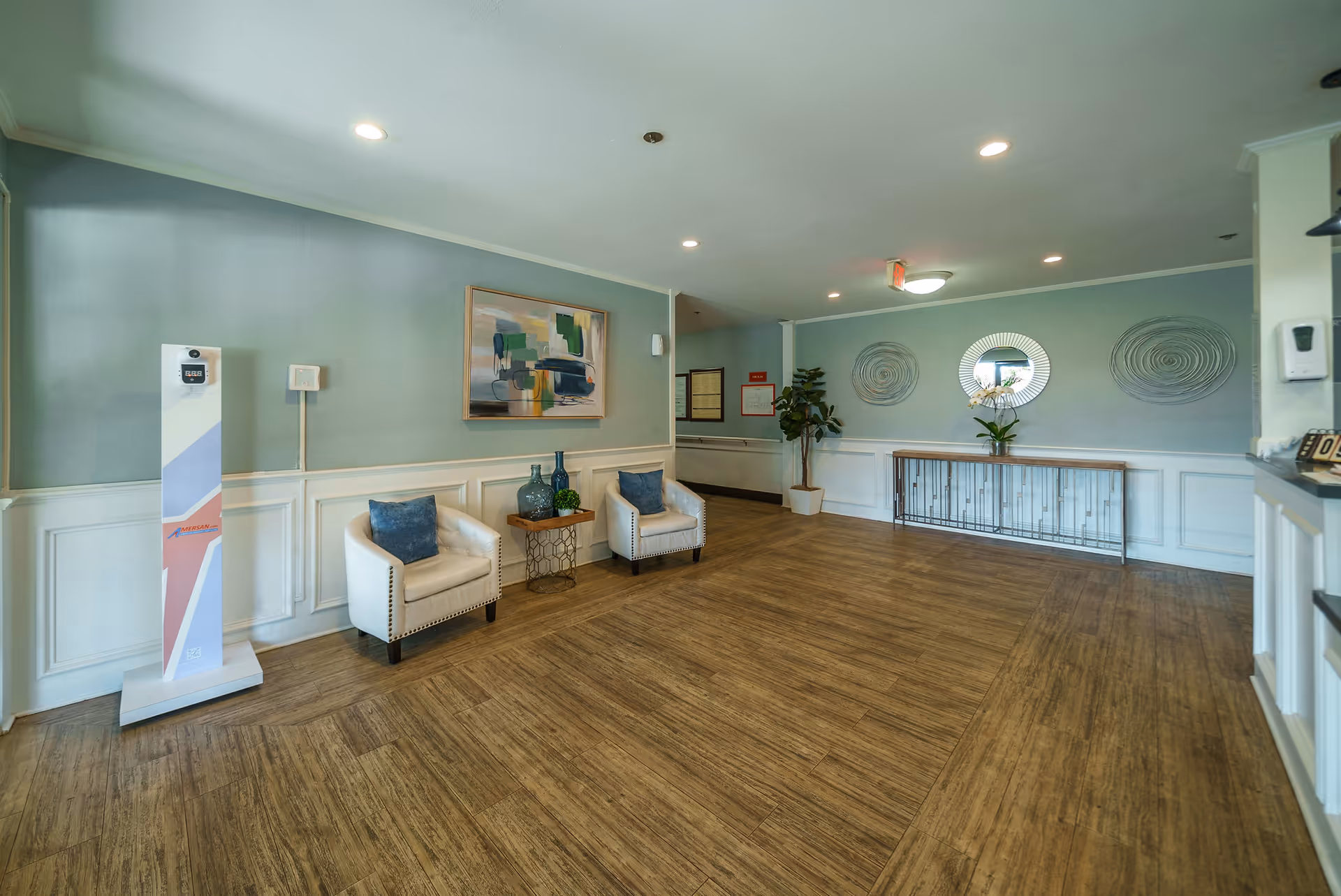A spacious interior room with wooden flooring and light green walls. The room features two white armchairs with blue cushions, a small wooden side table with decorative glass bottles, a large abstract painting on the wall, and a console table with a round mirror and decorative items. There is also a potted plant and a tall white device with a screen on the left side. The ceiling has recessed lighting.