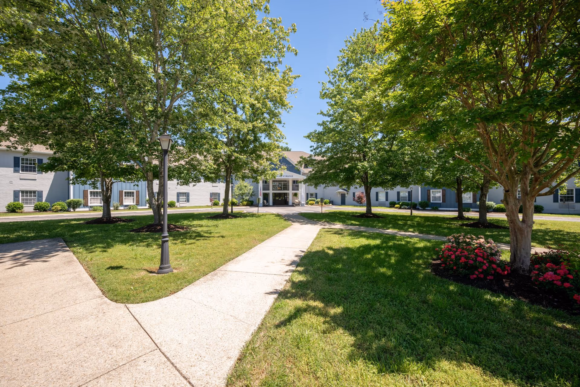 View of a senior living facility exterior with a paved walkway leading to the entrance. The building is surrounded by green grass, trees, and flower beds under a clear blue sky.