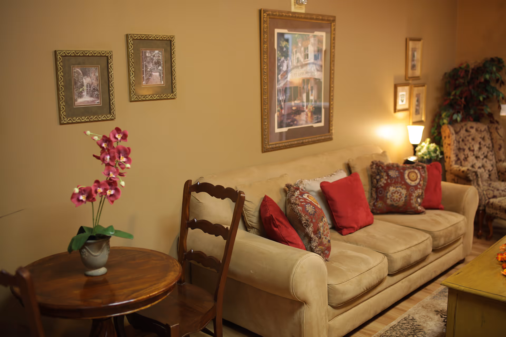 Cozy living room with a beige sofa adorned with red and patterned throw pillows, framed wall art, a small round table with pink orchids, and wooden chairs.
