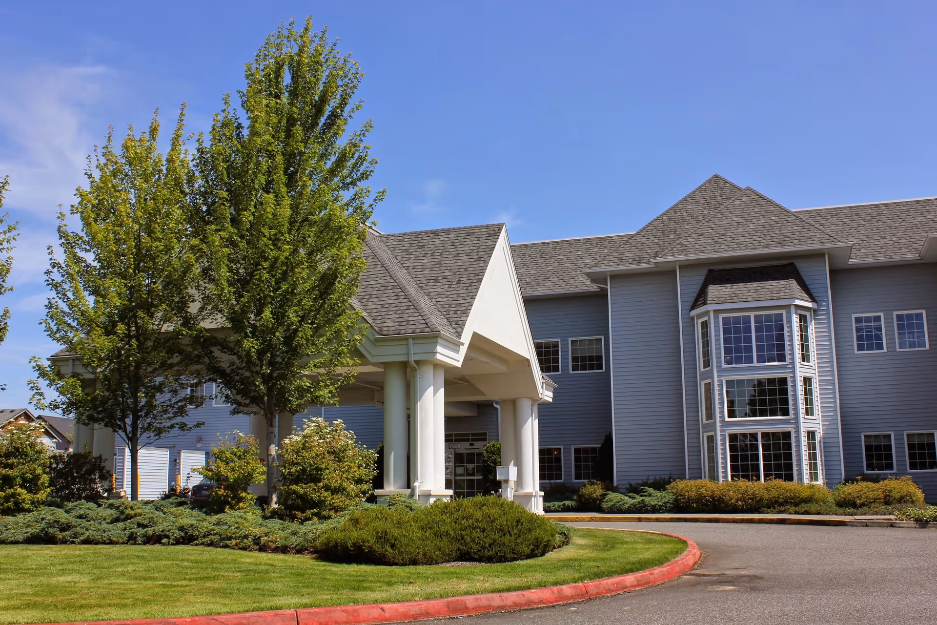 Exterior view of North Cascades Health and Rehabilitation building with a covered entrance, large windows, and well-maintained landscaping including trees and shrubs under a clear blue sky.