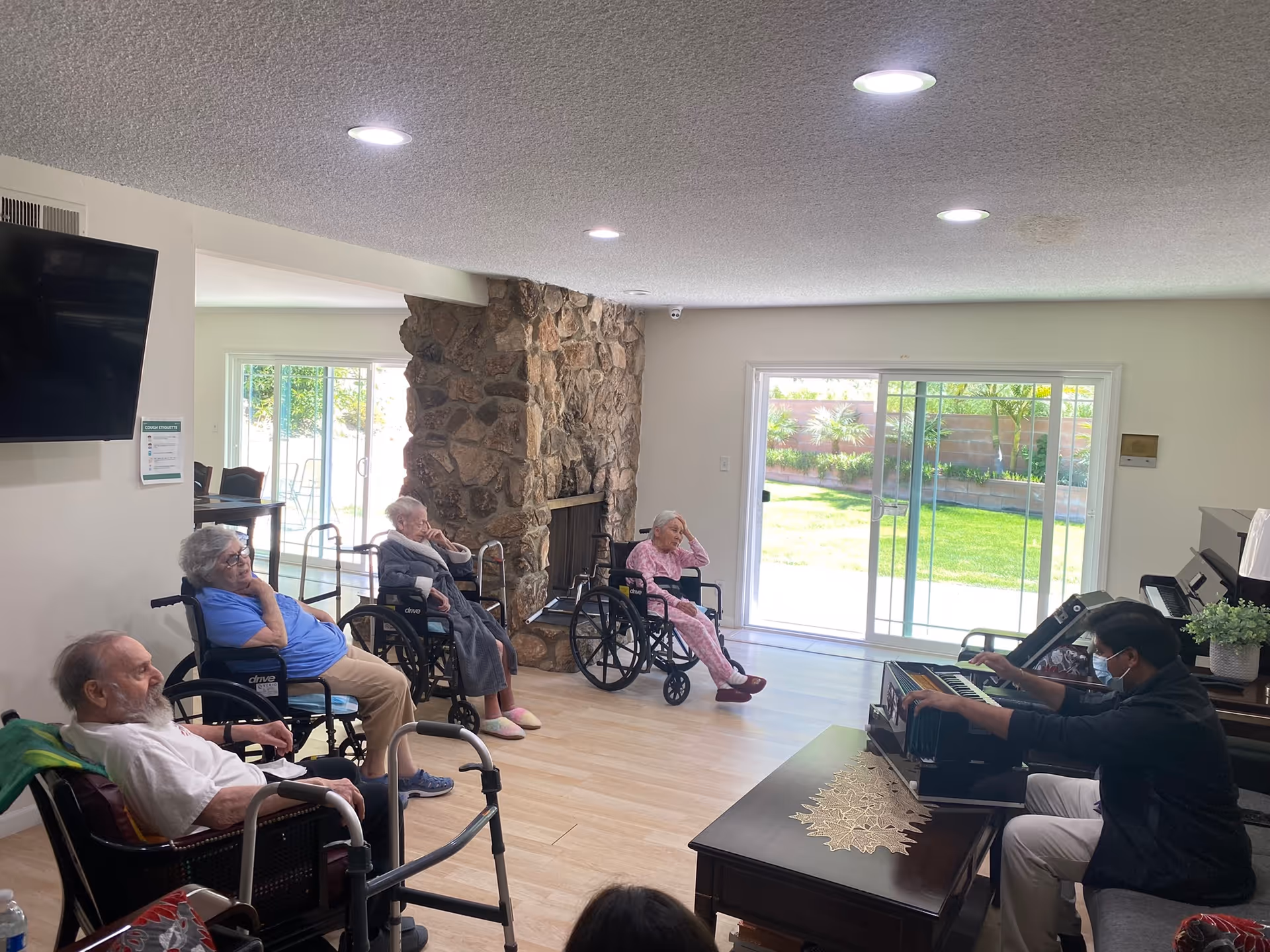 A group of elderly people sitting in wheelchairs in a living room area with a stone fireplace and large sliding glass doors leading to a garden. A man wearing a mask is playing a keyboard piano in front of them.