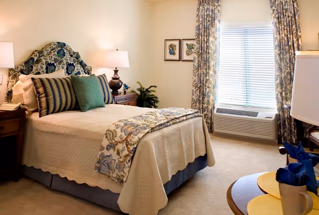 A cozy bedroom with a neatly made bed featuring a floral-patterned headboard and matching throw blanket. The bed has multiple pillows in green and striped patterns. There are two bedside tables with lamps, a window with floral curtains, and framed butterfly artwork on the wall. A small round table with a yellow placemat and a blue napkin is partially visible in the foreground.