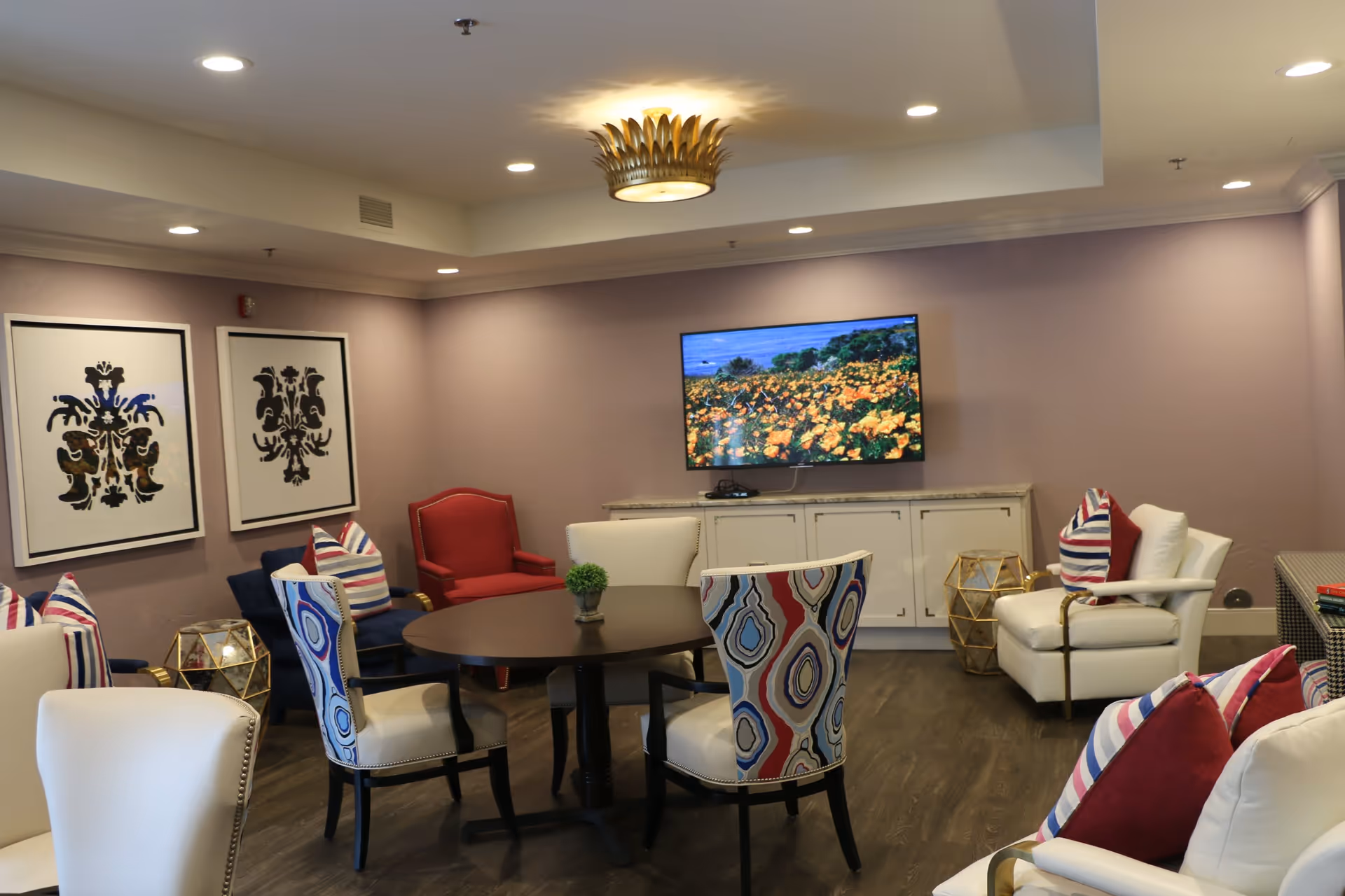 A decorated senior living room/lounge with colorful chairs and pillows around a round table, wall art, and a wall-mounted TV.