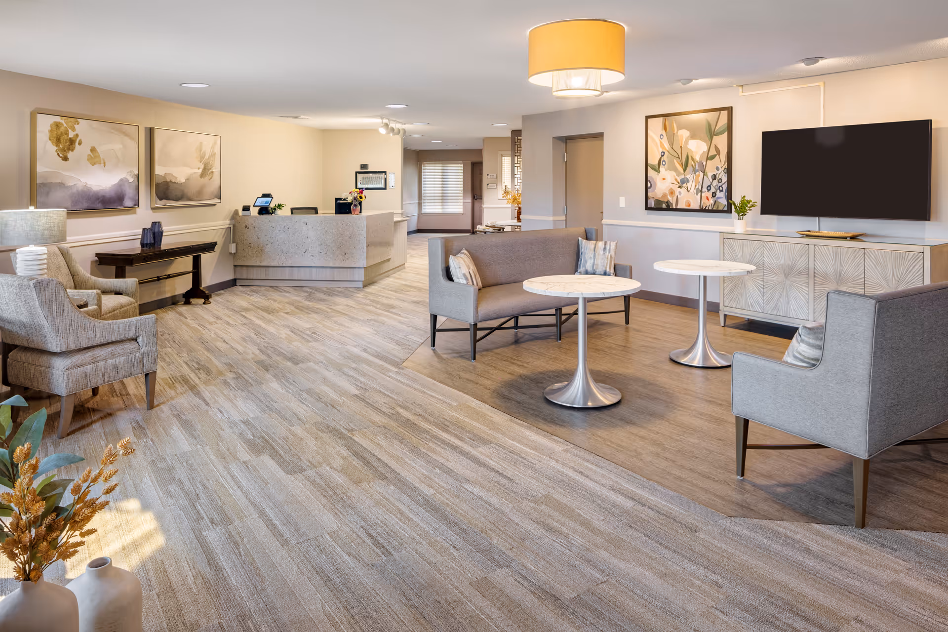 A spacious and well-lit senior living facility common area with a reception desk in the background. The room features comfortable seating including armchairs and a loveseat, two round tables, a large flat-screen TV mounted on the wall, and decorative artwork. The flooring is a combination of carpet and wood-like material, and there are modern light fixtures on the ceiling.