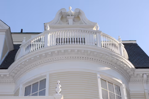 Close-up view of the upper exterior of a white mansion-style building with a curved balcony railing and decorative architectural details under a clear blue sky.