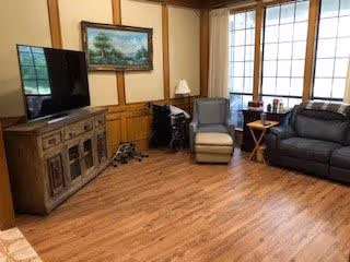A cozy living room with wooden flooring and wood-paneled walls. The room features a large flat-screen TV on a wooden cabinet, a wheelchair, a reclining chair, a small wooden side table with a lamp, and a dark leather sofa. Large windows with white blinds allow natural light to fill the space. A framed landscape painting hangs on the wall above the wheelchair.