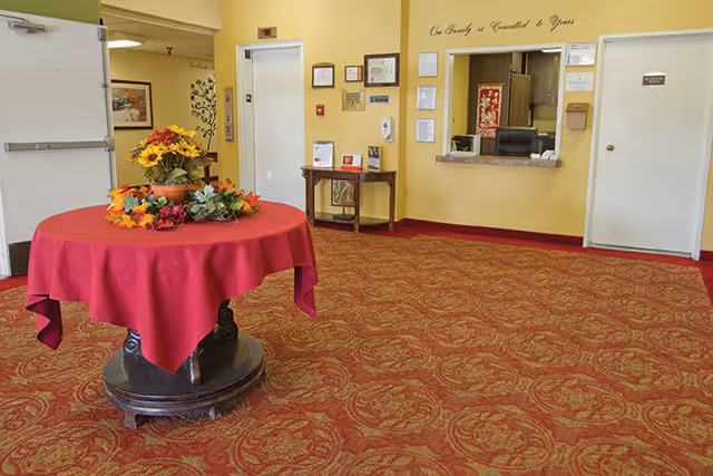 Interior view of a senior living facility lobby with a round table covered by a red tablecloth and decorated with a floral arrangement. The walls are light yellow, and there is a reception window with a chair behind it. Several framed certificates and notices are displayed on the wall. The carpet has a red and gold ornate pattern.