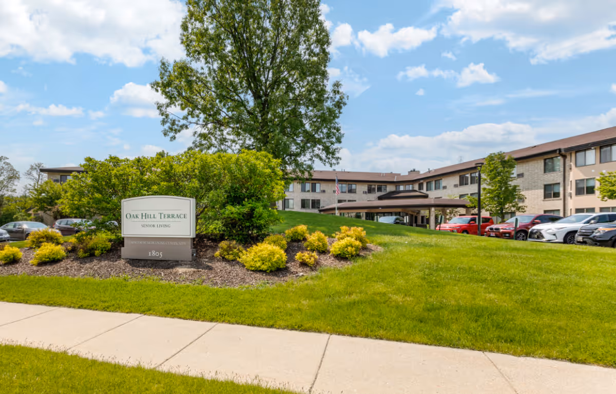 Exterior view of Oak Hill Terrace Senior Living facility with a landscaped garden, green lawn, and a large tree in front. The building is a multi-story structure with beige brick walls and multiple windows. Several cars are parked near the entrance under a covered driveway. The sky is partly cloudy with blue patches.
