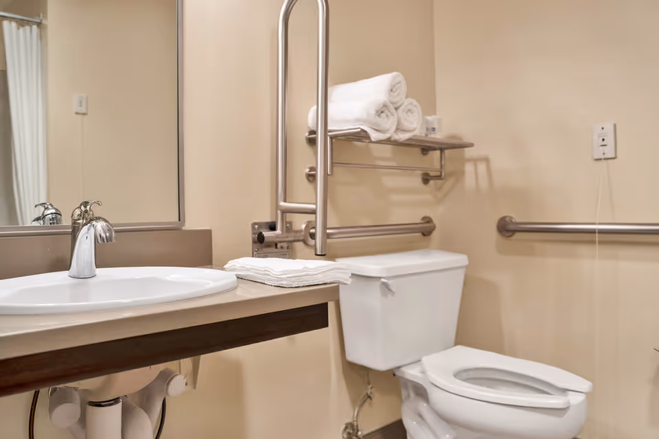 Accessible bathroom with a sink and mirror, toilet, grab bars, and folded towels on a shelf.