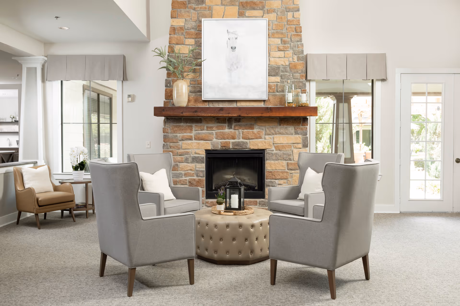 A cozy sitting area in an assisted living facility featuring four gray armchairs arranged around a tufted round ottoman with a decorative lantern on top. Behind the seating is a stone fireplace with a wooden mantel, adorned with a vase and candles. Above the mantel hangs a framed picture of a white horse. The room has large windows with beige valances and a glass door letting in natural light.
