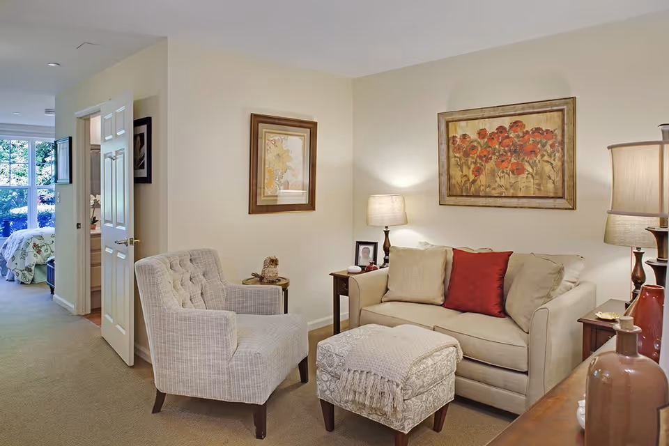 Cozy living room with a beige sofa, patterned armchair, ottoman, lamps, and framed floral artwork.