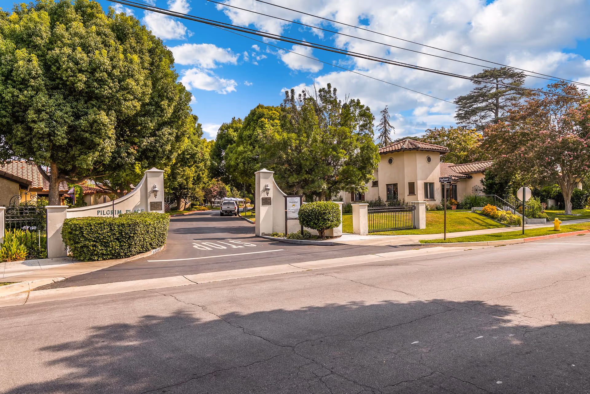 Entrance to Pilgrim Place Health Services Center with a gated driveway flanked by white stucco pillars and lush green trees. A street with a stop sign marking is visible in the foreground under a partly cloudy blue sky.