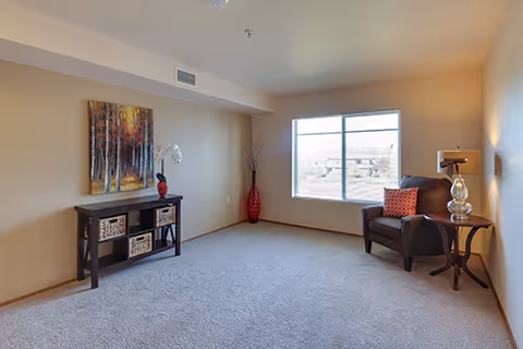 A cozy living room with beige walls and carpeted floor. There is a large window letting in natural light, a dark brown armchair with an orange patterned pillow, a small round side table with a glass lamp, a black console table with baskets and a red vase with white flowers, and a tall red decorative vase in the corner. A colorful abstract painting hangs above the console table.