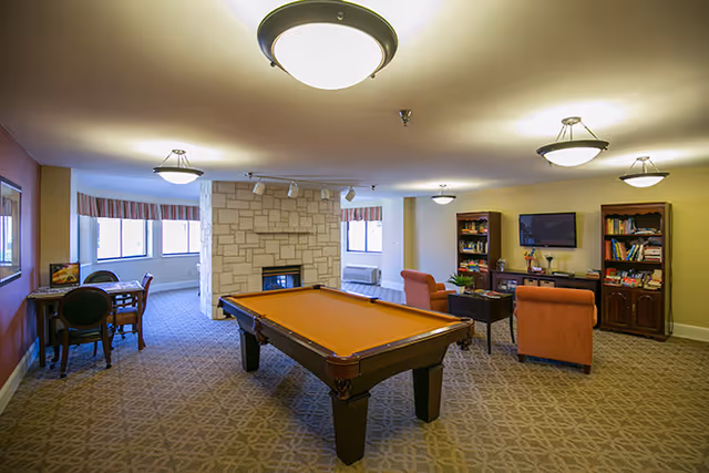 A spacious common area with a pool table in the center, two orange armchairs facing a TV mounted on the wall, bookshelves filled with books, a stone fireplace, and a small table with chairs near the windows.