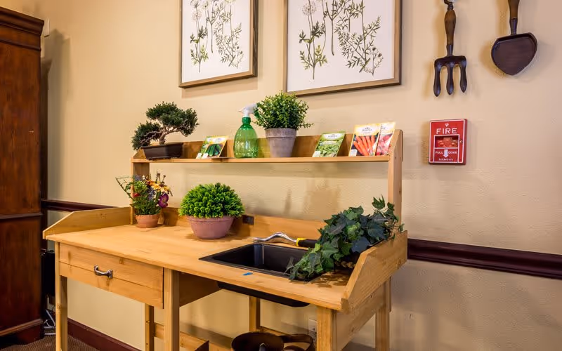 A wooden potting bench with a small sink, potted plants, seed packets and wall decorations in an indoor communal area.