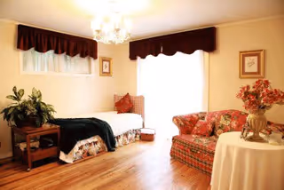A cozy living room area with a floral patterned sofa, a small table with a vase of flowers, a daybed with pillows and a blanket, hardwood floors, and windows with dark valances letting in natural light.