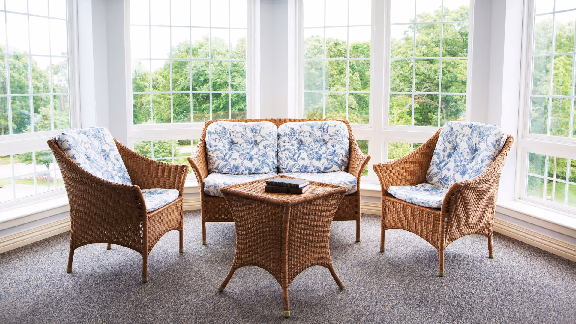 A bright sunroom with large windows overlooking green trees, furnished with a wicker loveseat and two wicker armchairs with blue and white floral cushions, and a matching wicker coffee table with two books on top.