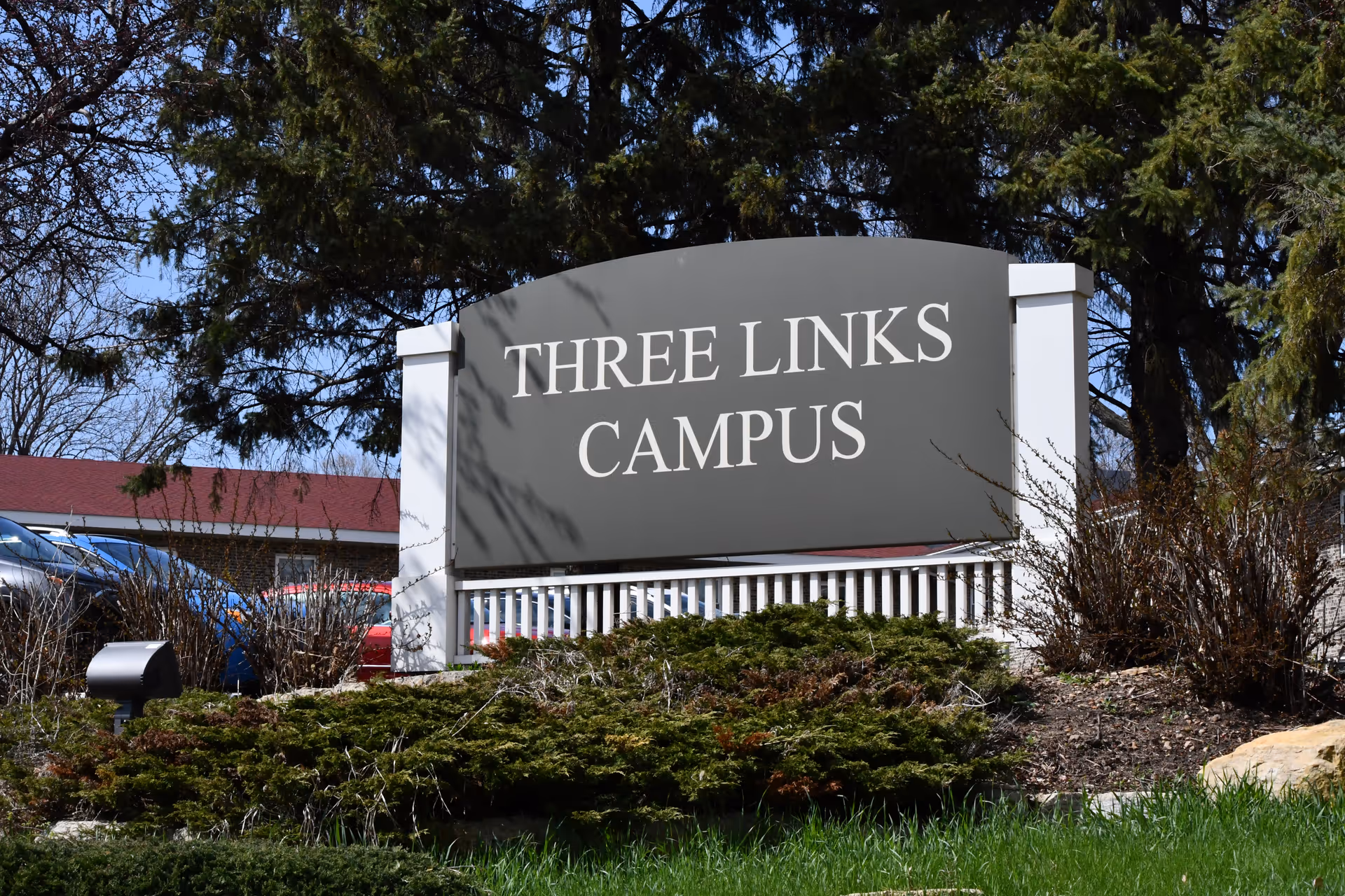 Outdoor view of a large sign that reads 'THREE LINKS CAMPUS' surrounded by greenery and trees, with a building and parked cars visible in the background under a clear blue sky.