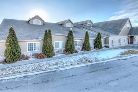 Front exterior of a single-story assisted living building with evergreen trees and light snow.