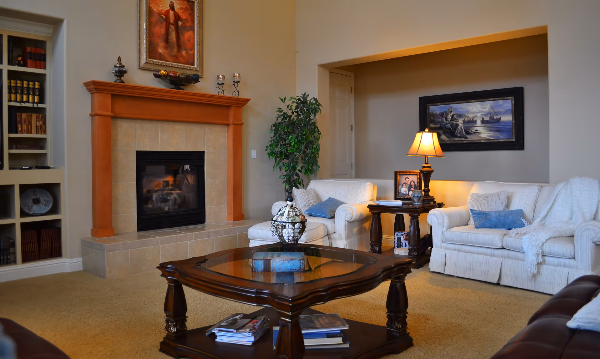 A cozy living room with a beige carpet, a fireplace with a wooden mantle, two white upholstered sofas with blue and beige cushions, a wooden coffee table with books and magazines, a side table with a lamp and framed photos, a green potted plant, and artwork on the walls.