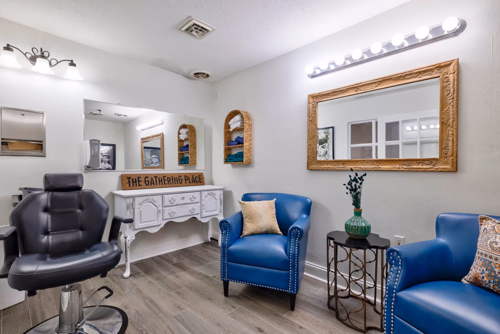 Interior room with a black salon chair, two blue armchairs with decorative pillows, a small black side table with a green vase and flowers, a large ornate mirror with lights above it, a white vintage-style dresser with a wooden sign that reads 'THE GATHERING PLACE', and shelves with folded towels on the wall.