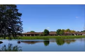 A serene outdoor scene showing a calm pond with clear reflections of trees and a low building in the background under a blue sky.