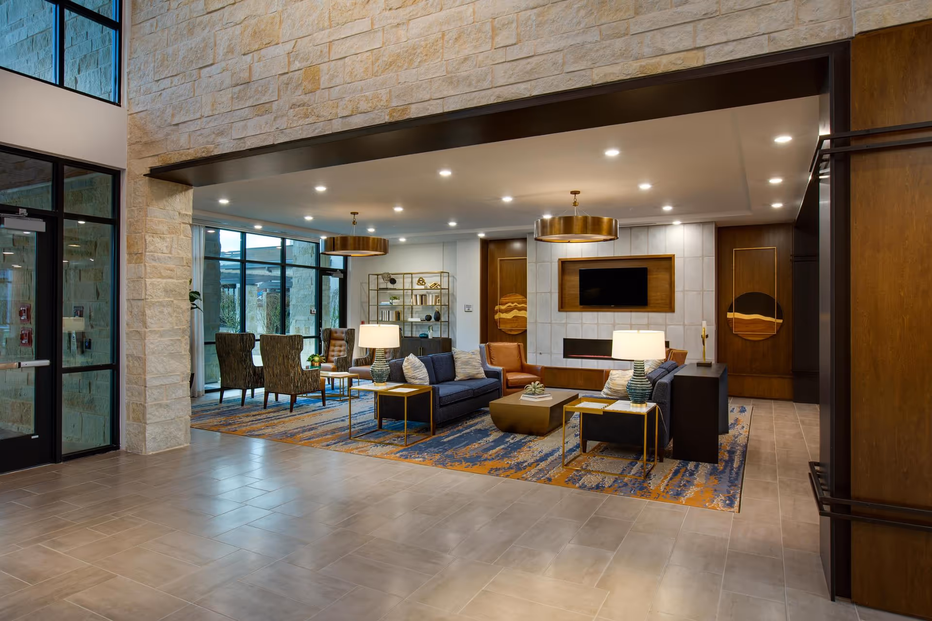 A modern and spacious living room area with a large patterned rug, two blue sofas, two brown armchairs, side tables with lamps, and a wall-mounted flat screen TV above a built-in fireplace. The room features stone walls, large windows, and decorative lighting fixtures hanging from the ceiling.