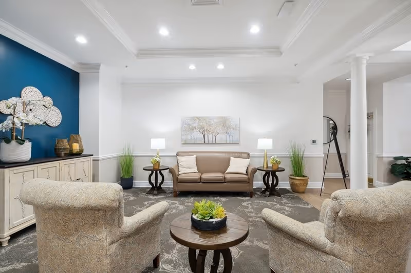 A well-lit living room with a beige leather sofa against a white wall, flanked by two side tables each with a lamp and small plant. Two patterned armchairs face the sofa with a round wooden coffee table with a small plant in the center. A white cabinet with decorative items and a blue accent wall is on the left side. The room has a patterned carpet, white columns, and a ceiling with recessed lighting.