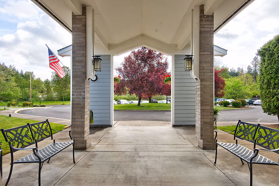 Covered entrance area of a facility with two black metal benches facing each other, brick pillars supporting the roof, an American flag on the left side, and a view of a parking lot and trees with green and red foliage in the background.