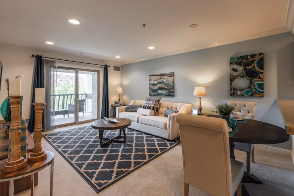 A cozy living room with a beige sofa adorned with patterned cushions, a round wooden coffee table on a dark patterned rug, two table lamps on side tables, and abstract wall art. There is a sliding glass door leading to a balcony with outdoor seating, and a small dining table with chairs is visible in the foreground.