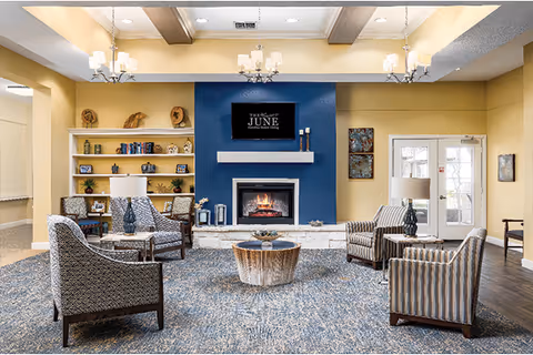 A cozy living room area in a senior living facility with four upholstered armchairs arranged around a round wooden coffee table. The room features a blue accent wall with a lit fireplace and a mounted TV above it displaying 'The Waterford at June'. There are built-in shelves with decorative items and books on the left side, two chandeliers hanging from the ceiling, and a door with windows on the right side letting in natural light.
