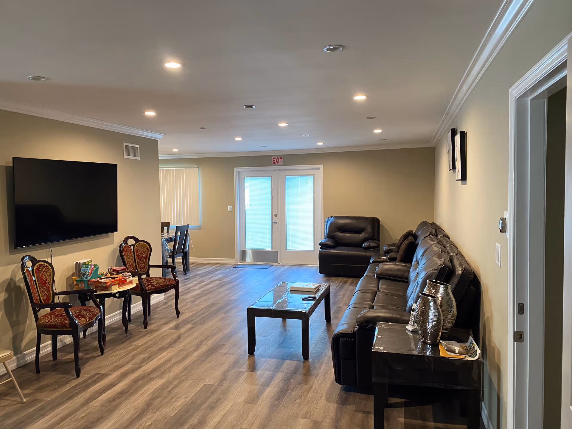 A spacious living room with wood flooring, beige walls, and recessed ceiling lights. The room features a large black leather sofa, a matching armchair, a glass coffee table, and two ornate chairs with red and gold upholstery. A flat-screen TV is mounted on the wall, and a small table with board games is placed between the chairs. In the background, there is a dining area with a table and chairs near a window with vertical blinds, and a set of double glass doors with an exit sign above them.
