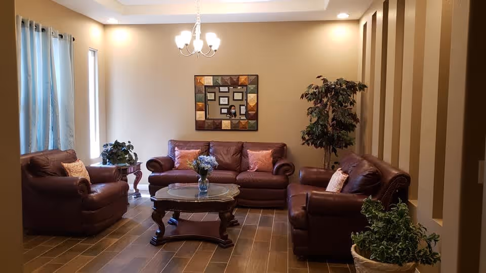 A cozy living room with three brown leather sofas arranged around a wooden coffee table with a vase of flowers. There are decorative pillows on the sofas, a large framed mirror on the wall, a chandelier hanging from the ceiling, and several potted plants placed around the room. Light blue curtains cover a window on the left side.