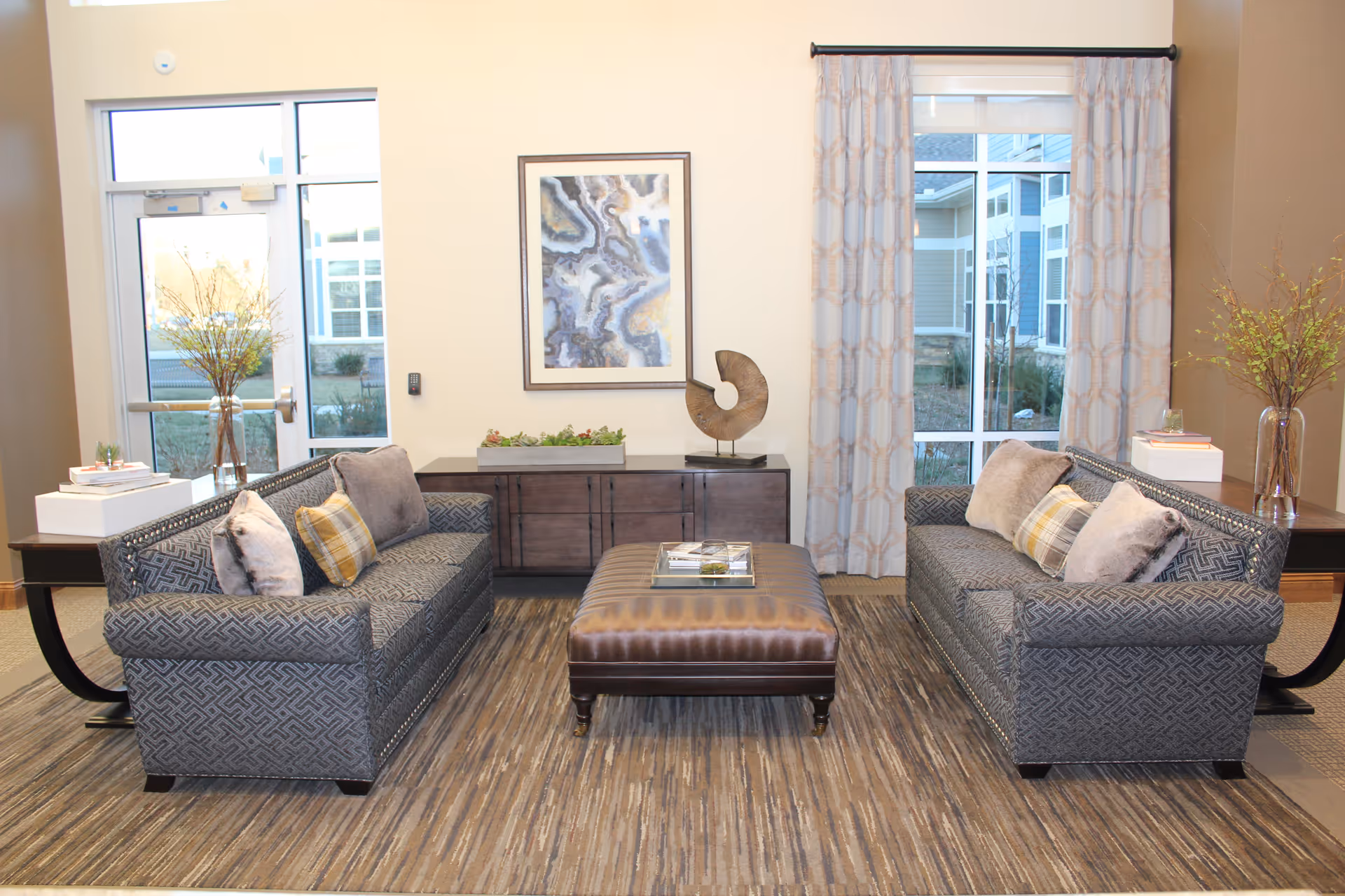 A well-lit living room with two patterned gray sofas facing each other, each adorned with multiple cushions including plaid and solid colors. Between the sofas is a large brown leather ottoman with books on top. Behind the ottoman is a dark wooden sideboard with decorative items including a sculpture and a planter. The room has large windows with patterned curtains, and there are vases with yellow flowers on side tables next to each sofa. A framed abstract artwork hangs on the wall above the sideboard.