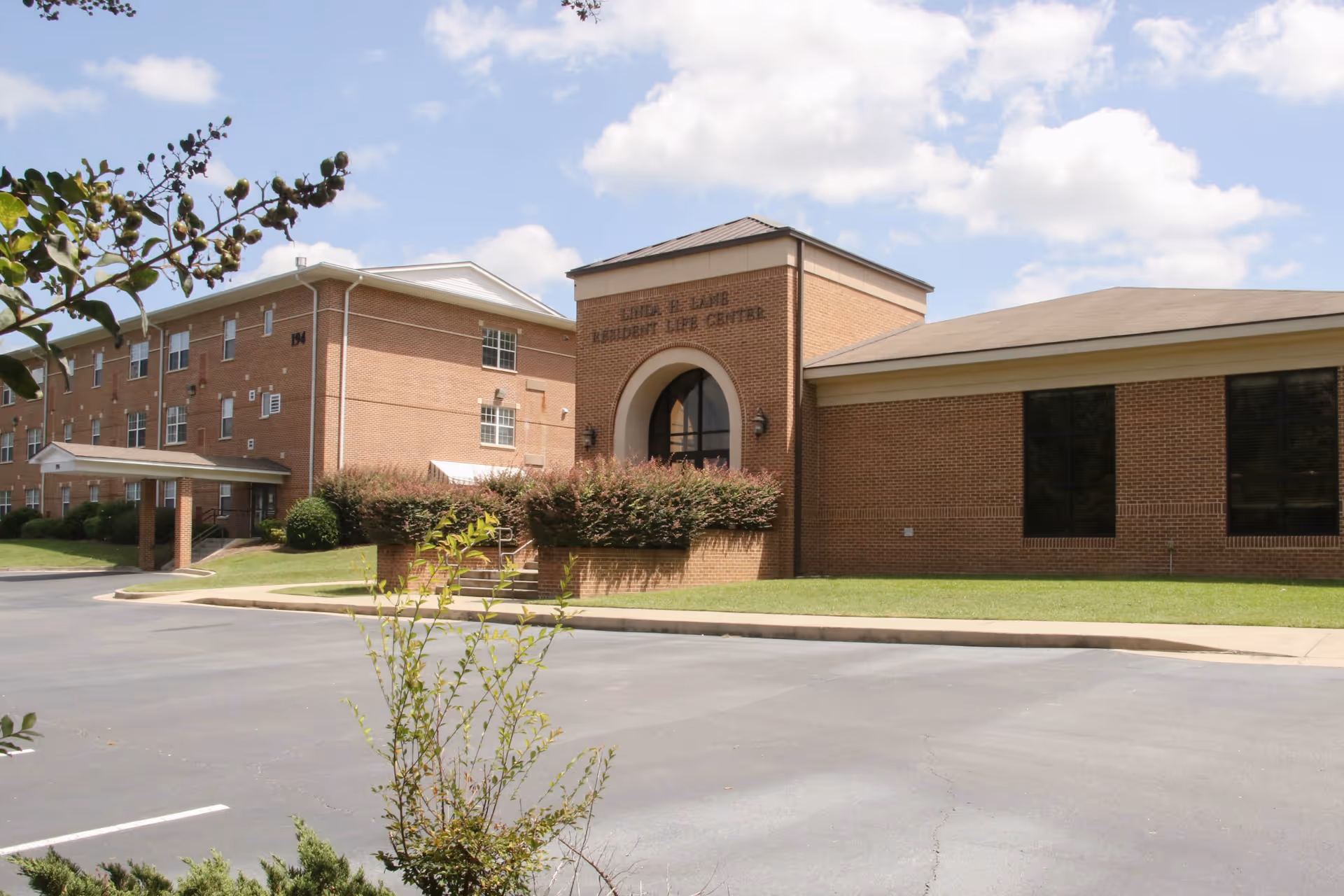 Exterior view of a brick building complex under a partly cloudy sky. The building has multiple windows and a covered entrance. There is a parking lot in front with some greenery and small trees near the sidewalk.