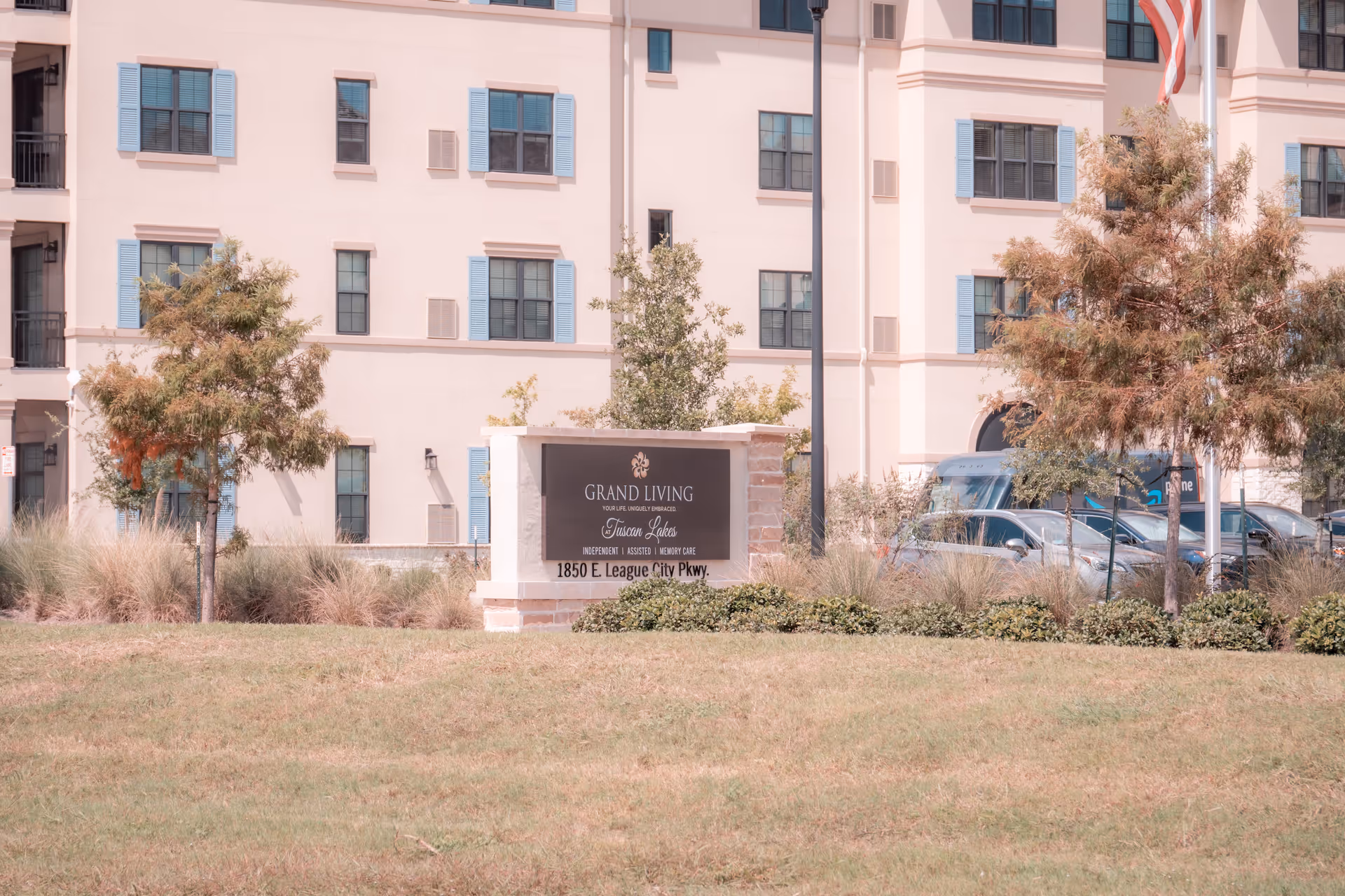 Exterior view of the Grand Living At Tuscan Lakes senior living facility with a sign displaying the facility name and address in front of a multi-story building with blue window shutters, trees, and parked cars.
