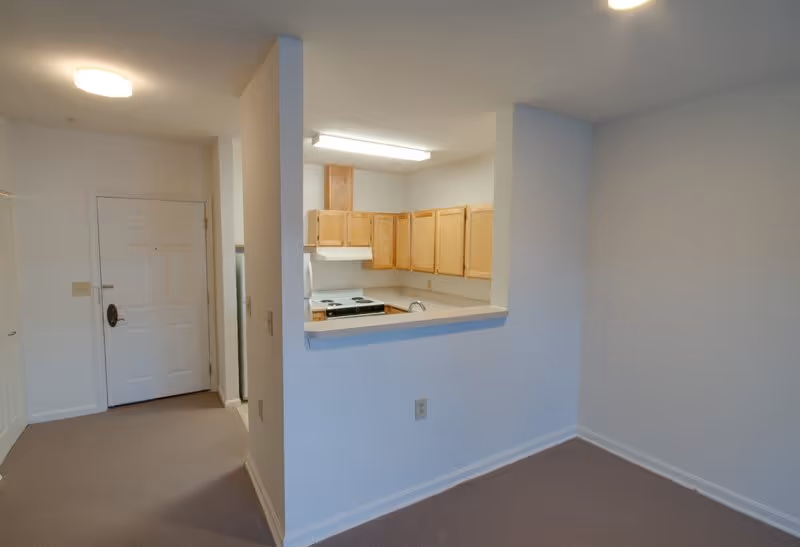 Interior view of a small apartment or senior living unit showing an entry door, a kitchen with wooden cabinets, a stove, and a refrigerator partially visible behind a half wall, and an adjacent empty carpeted area with white walls and ceiling lights.