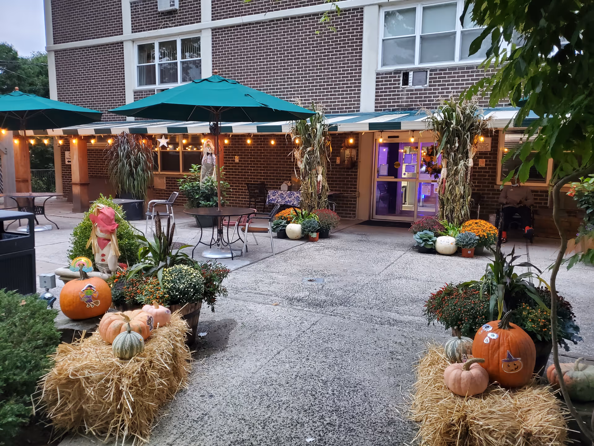 Outdoor courtyard area of a senior living facility decorated for fall with pumpkins, hay bales, potted plants, and string lights. There are green umbrellas over tables and chairs, and the building entrance is visible in the background with autumn decorations around the door.