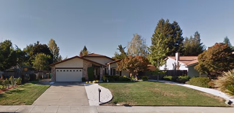 Front exterior view of a single-story residential house with a two-car garage, a driveway, a mailbox, and a well-maintained lawn with trees and shrubs under a clear sky.