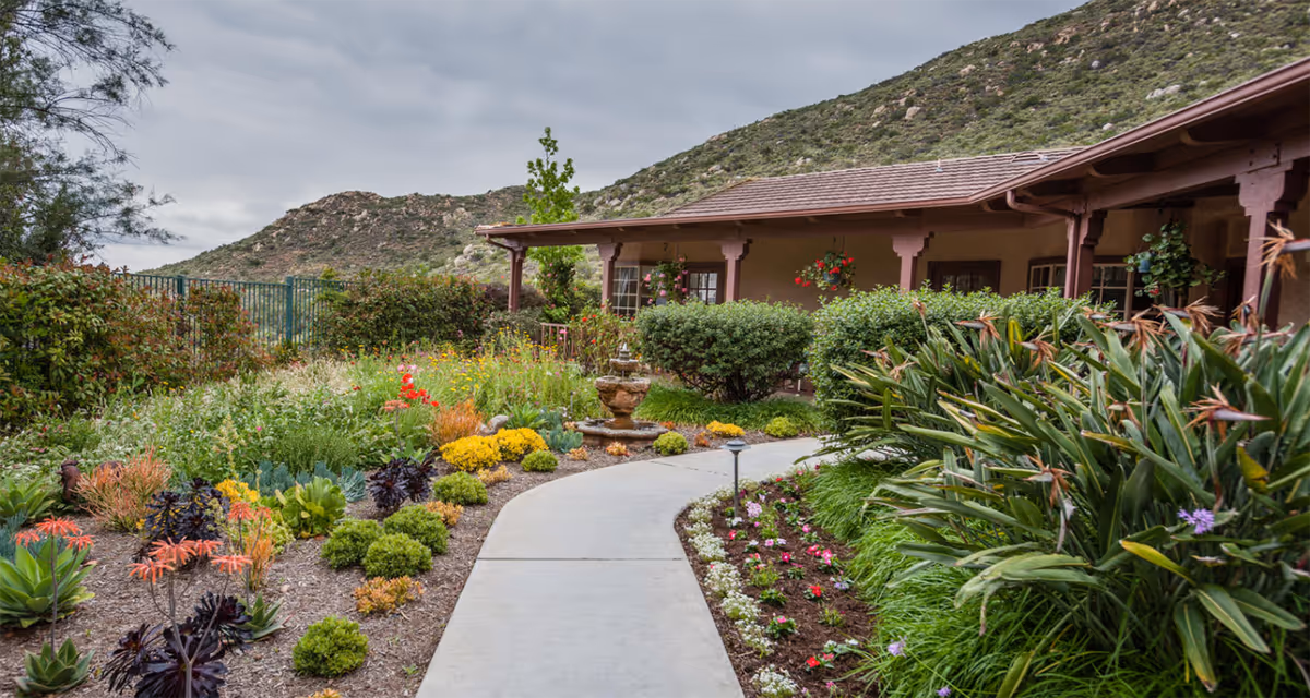 Curved concrete path through a colorful landscaped garden leading to a covered porch of a single-story building with hills in the background.