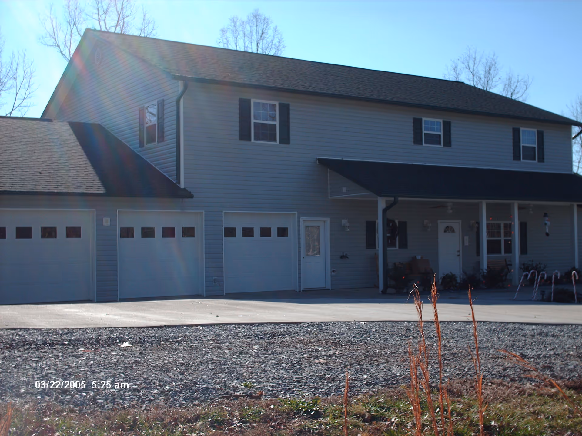 Exterior view of a two-story building with gray siding and a black roof. The building has three garage doors on the left side and a covered porch area on the right with a white door and windows. There are some plants and candy cane decorations near the porch. The sky is clear and the sun is shining, creating lens flare in the image.