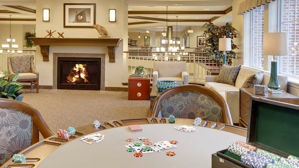 A cozy senior living common area with a poker table in the foreground featuring playing cards and poker chips. In the background, there is a lit fireplace with a mantel decorated with small sculptures and a framed picture above it. Comfortable chairs and sofas are arranged around the room, with lamps and plants adding to the warm ambiance. Large windows let in natural light.