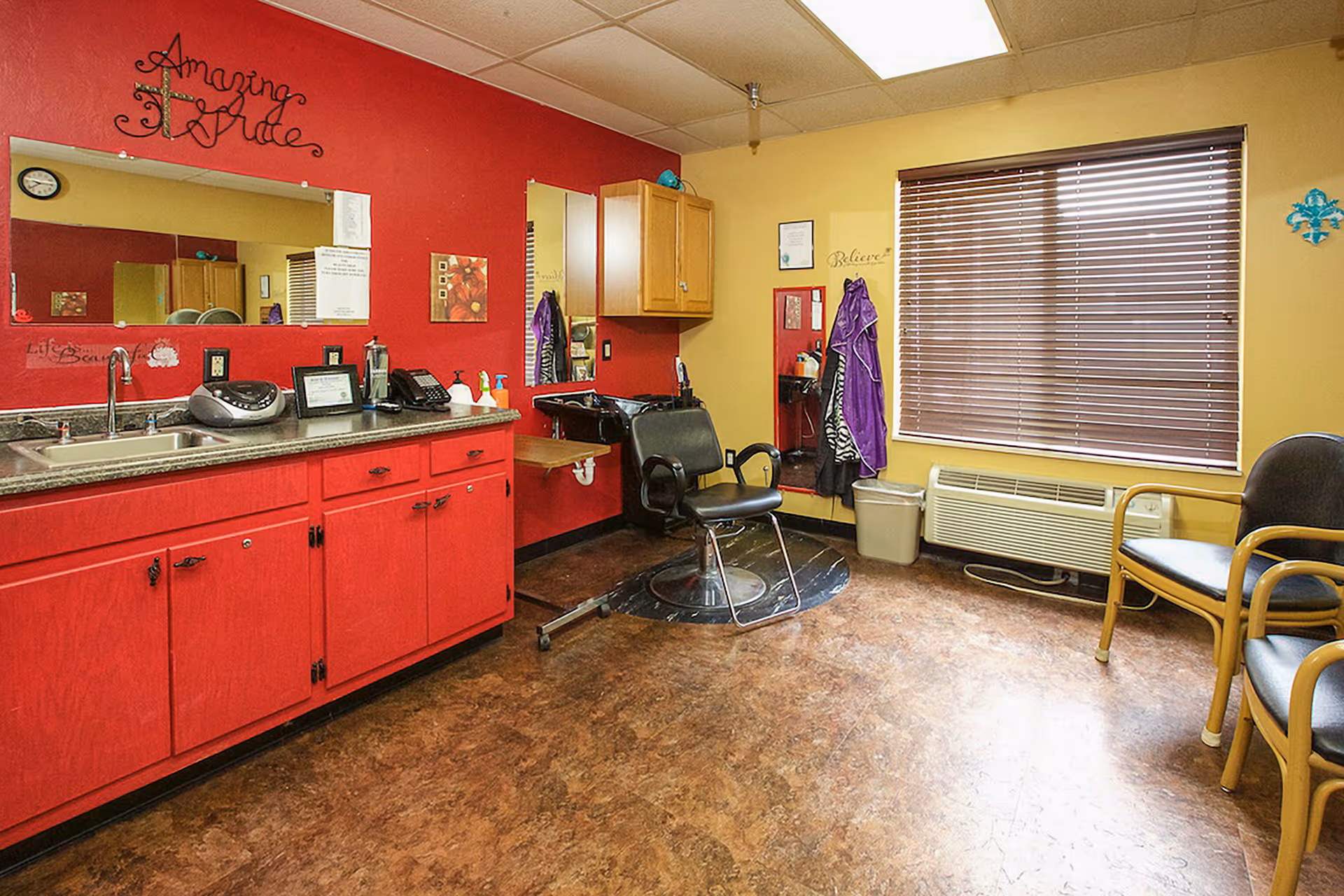 Interior of a salon area in a senior living facility with a red cabinet and countertop on the left, a black salon chair in front of a mirror on the right wall, a window with closed blinds, and two waiting chairs with wooden armrests on the right side.