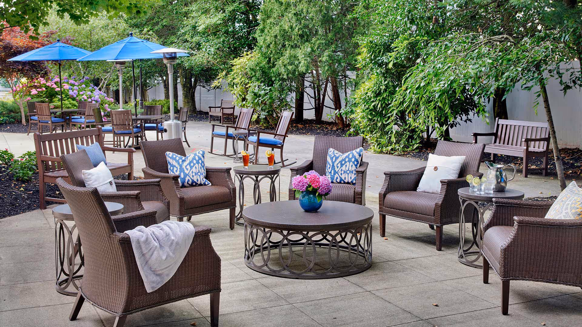 Outdoor patio area with brown wicker chairs and sofas arranged around a circular metal coffee table with a blue vase of pink flowers. There are side tables with drinks and a pitcher, blue umbrellas shading tables with wooden chairs, and lush green trees and plants surrounding the space.