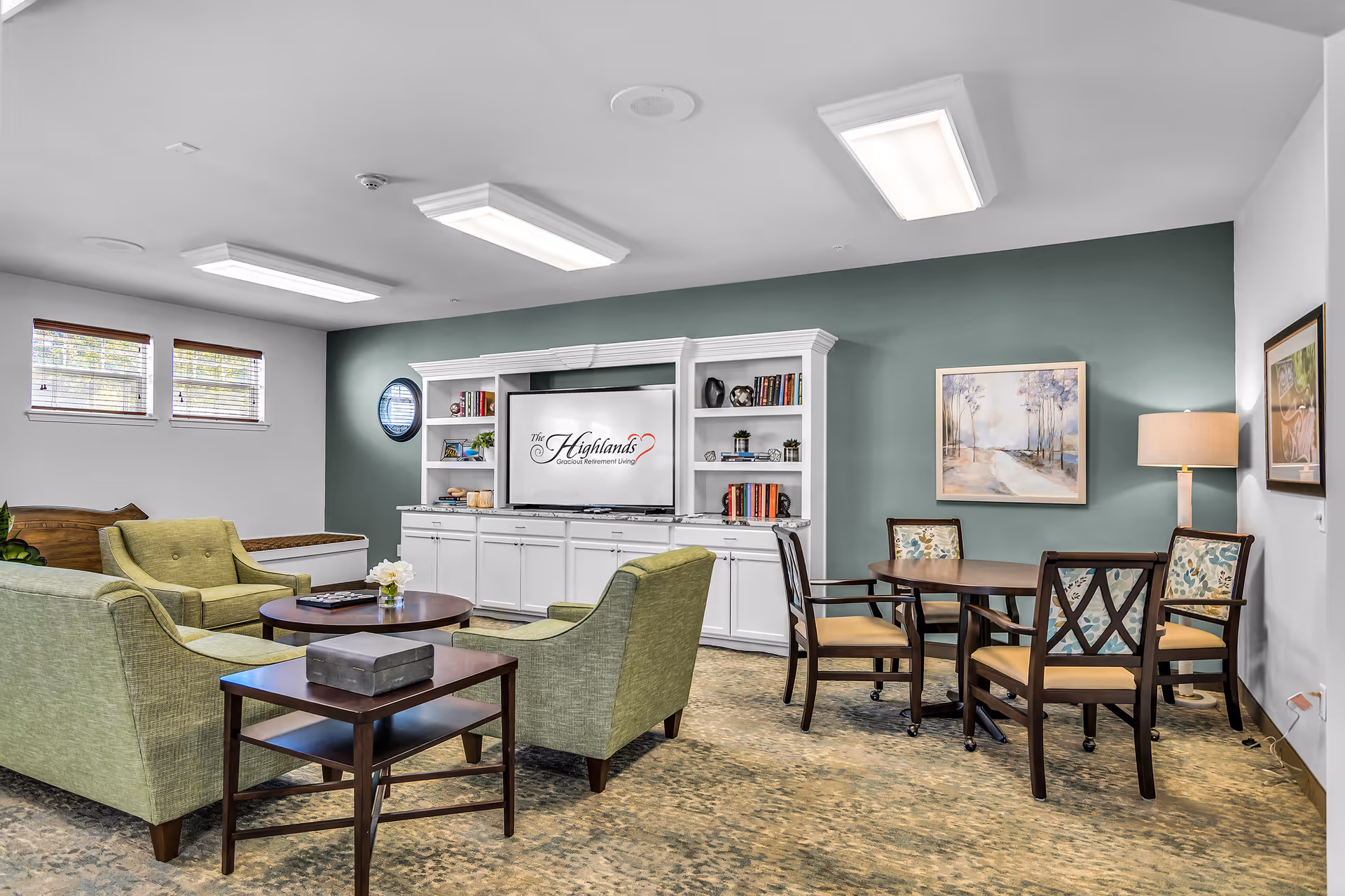 A cozy living room area in The Highlands Gracious Retirement Living featuring green upholstered armchairs and a sofa arranged around a round wooden coffee table. There is a white built-in entertainment center with shelves holding books and decorative items, and a TV displaying the facility's logo. To the right, there is a round wooden table with four chairs, a floor lamp, and framed artwork on the walls. The room has a green accent wall and carpeted floor.