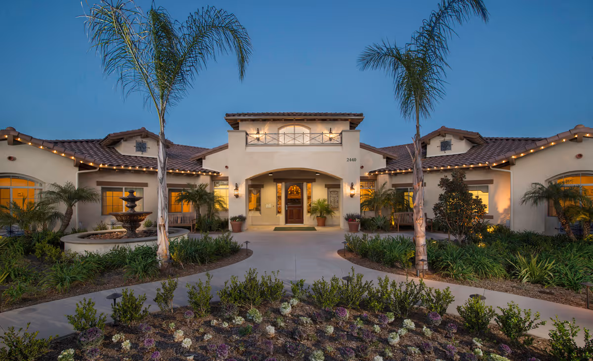 Front exterior view of ActivCare Mission Bay facility at dusk with a pathway leading to the entrance, flanked by palm trees, landscaped gardens, and a water fountain on the left side.