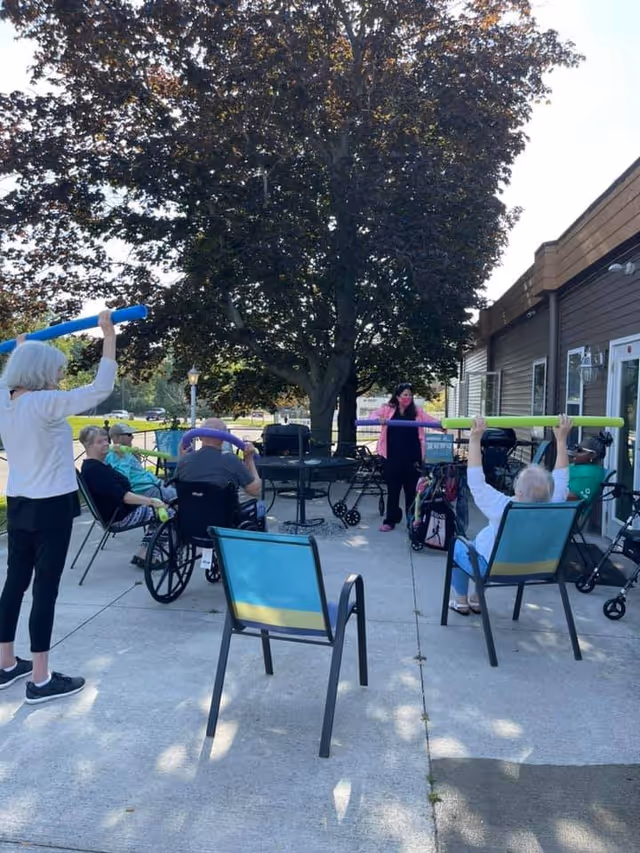 Several elderly residents and a staff member exercising with colorful foam noodles on an outdoor patio under a large tree.