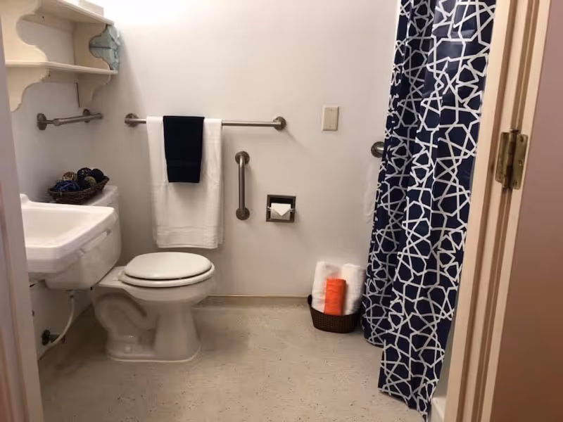 Small bathroom with a toilet, wall-mounted sink, grab bars, towel rack, and a blue patterned shower curtain with a basket of towels.