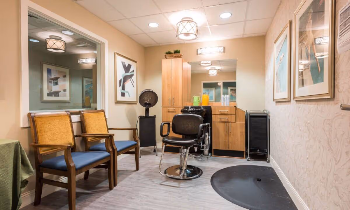 Interior view of a salon area in a senior living facility with two wooden chairs with yellow backs and blue seats, a black salon chair in front of a large mirror, wooden cabinets, a hair dryer, and framed artwork on the walls.