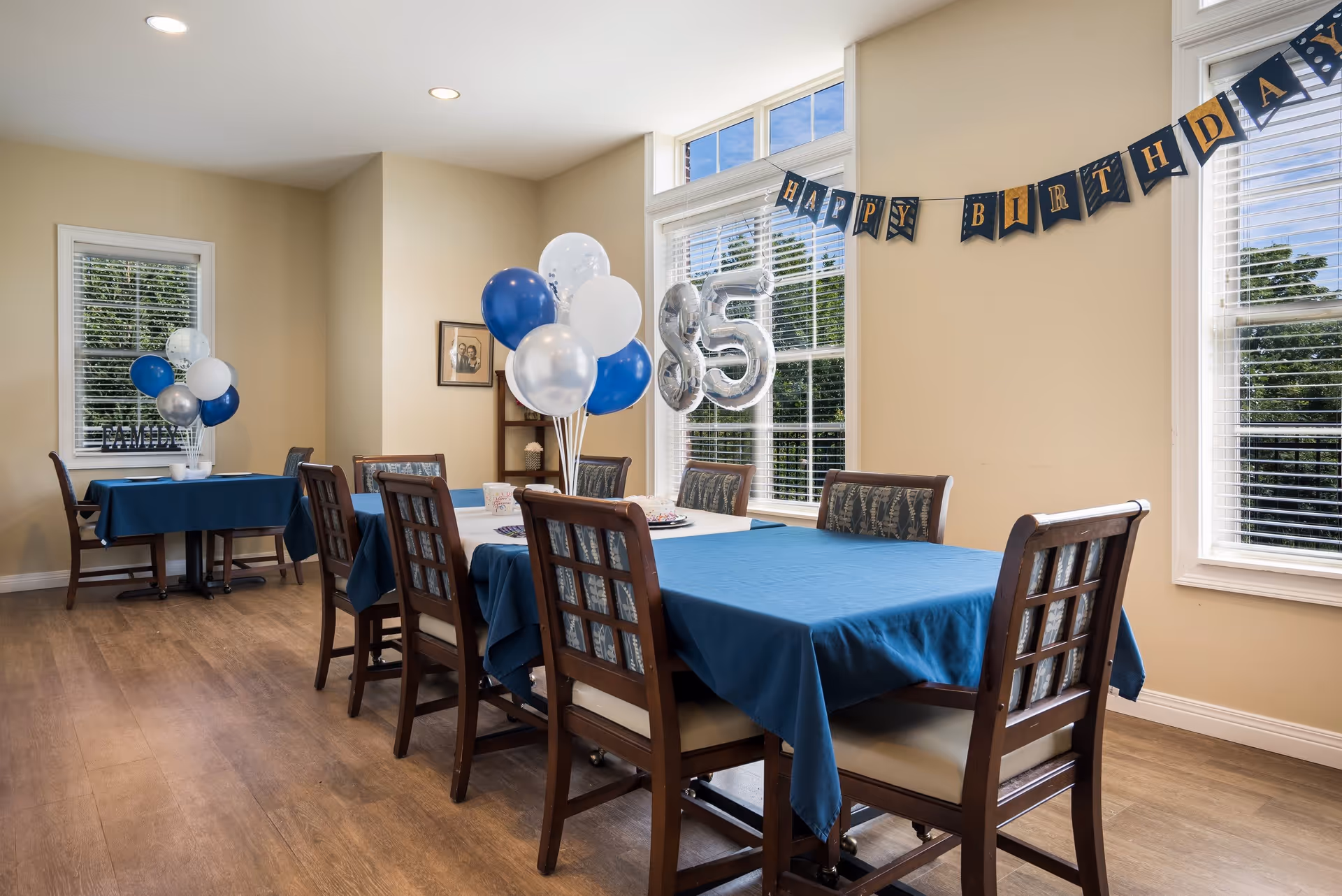 A dining room decorated for a birthday celebration with blue and white balloons, a 'Happy Birthday' banner, and silver balloons shaped as the number 85. The room has wooden floors, beige walls, and large windows with white blinds letting in natural light. Tables are covered with blue tablecloths and surrounded by wooden chairs with patterned cushions.